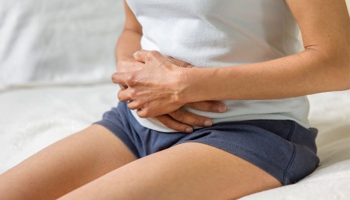A woman sitting on the side of her bed holding her stomach.