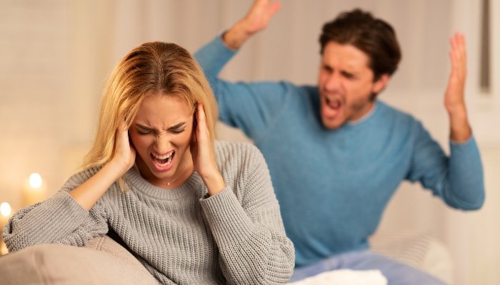 An angry man shouting at a woman with her hands over her ears while they sit on a couch.