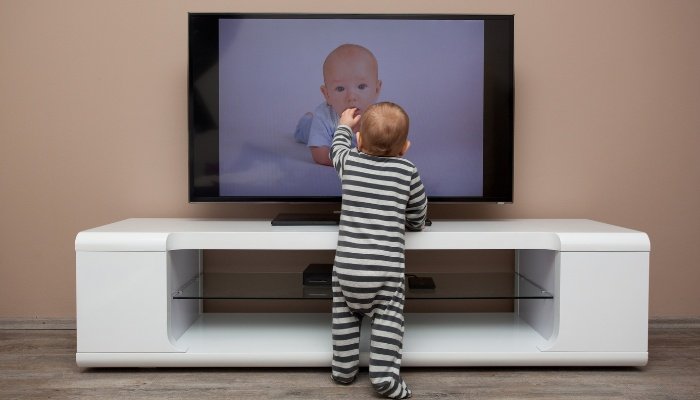 A baby boy standing in front of the tV touching the image of a baby on the screen.
