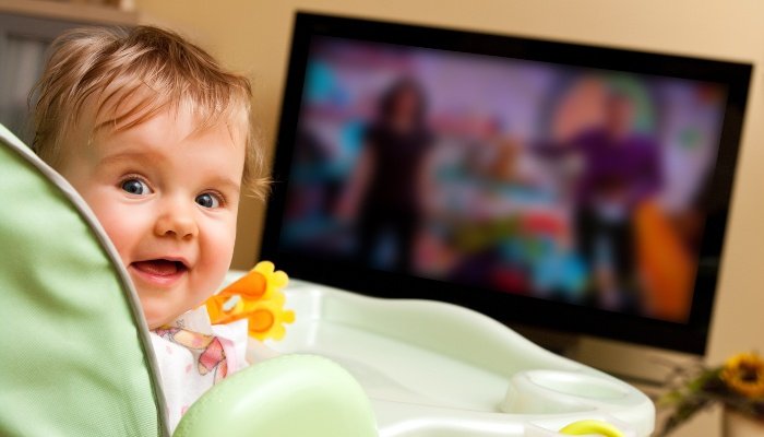 A baby girl sitting in her high chair watching TV.
