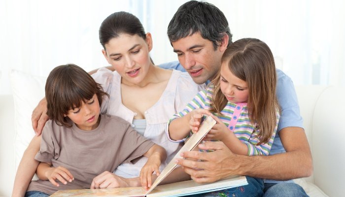 A family sitting on a white couch reading a book together.