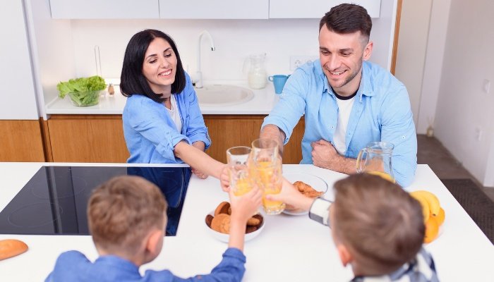 A man, woman, and two boys toast with glasses of orange juice at a kitchen counter.
