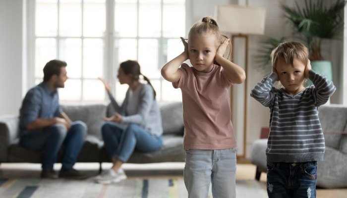 A brother and sister with their hands over their ears to block out parents' arguement.