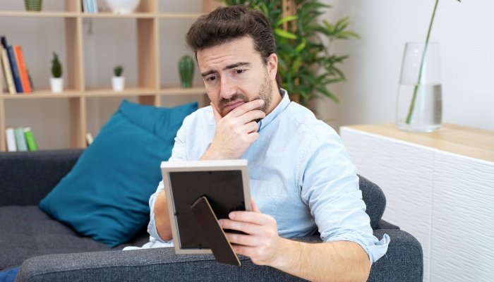 A lonely man sits on a couch while staring at a framed photo.