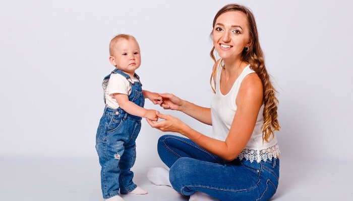A mother sitting on the floor helping her baby stand up.
