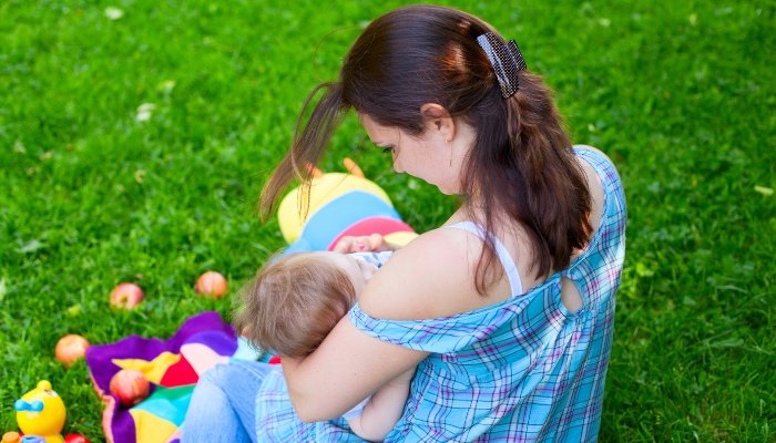 A young mother nursing her baby outside while sitting on a lush lawn surrounded by baby toys.