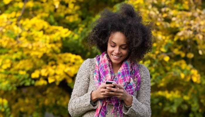 A smiling young woman reading a text message on her phone while standing outside on a pretty day.