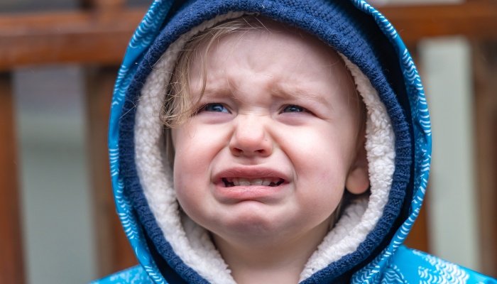 A three-year-old boy has a tantrum outside while wearing a blue raincoat.