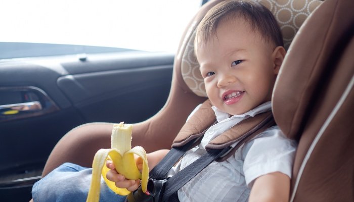 A cute toddler snacking on a banana while sitting in his car seat.