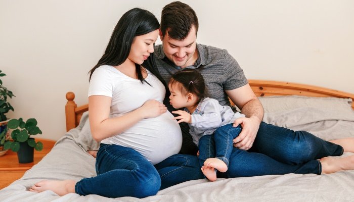 A family sitting on the bed with the toddler kissing the mother's pregnant belly.