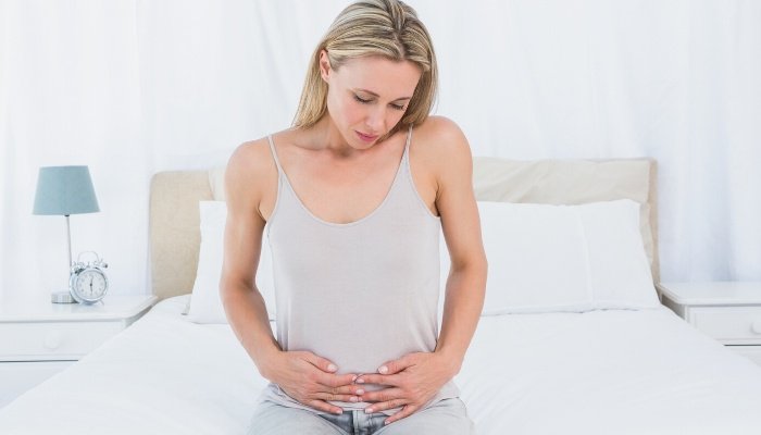 A young woman sitting on the side of her bed holding her lower abdomen and feeling unwell.