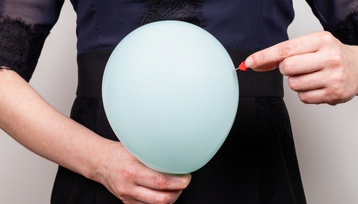 A woman holding a balloon in front of her stomach with a pin to pop it in her other hand.