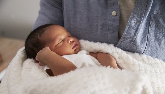 A father holding his newborn daughter who is loosely wrapped in a soft, white blanket.