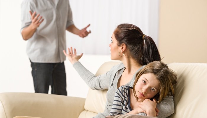 A man arguing with or yelling at a frustrated woman sitting on the couch holding her young daughter.