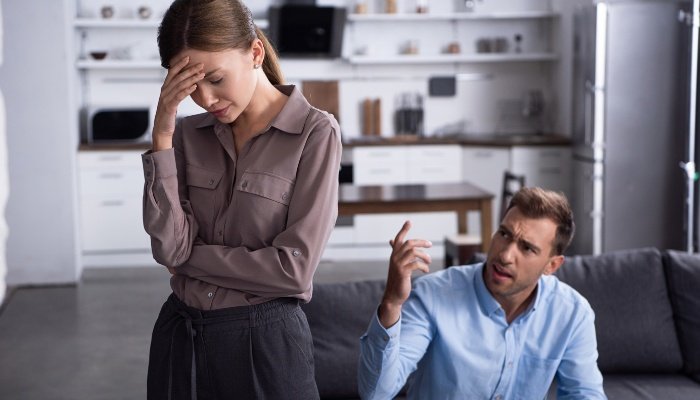A man sitting on the couch yelling at a woman standing and holding her head in frustration.