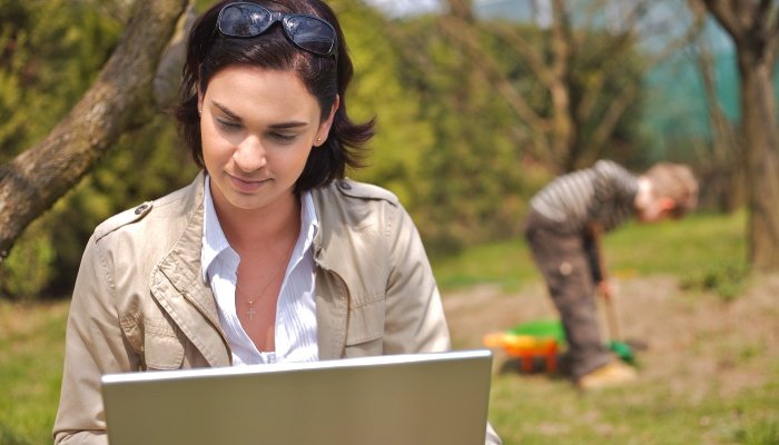 A young mother works on her laptop outdoors while her son plays in the background.