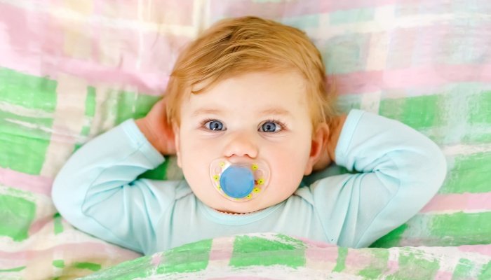 A red-headed toddler lies wide awake in her bed with a pacifier in her mouth.