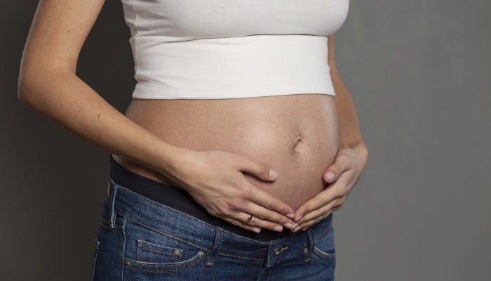 A pregnant lady with her hands on her belly against a gray background.
