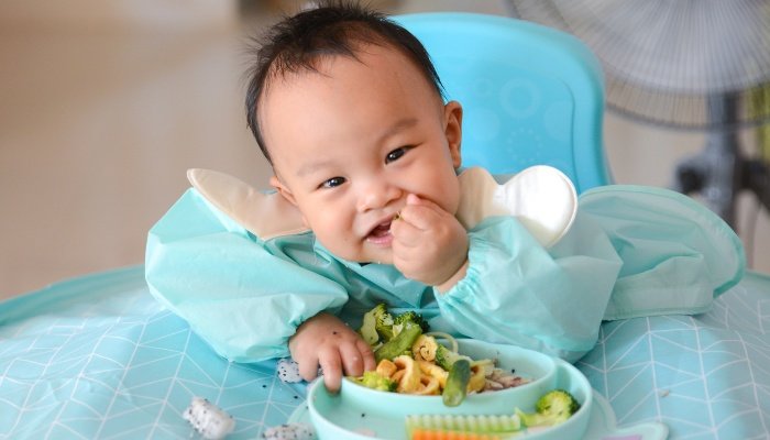 A cute baby boy sitting in a highchair with a smock bib feeding himself from a bowl.