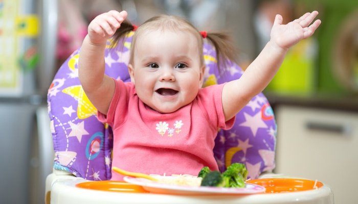 A cute baby girl in pigtails eating broccoli while sitting in a highchair.
