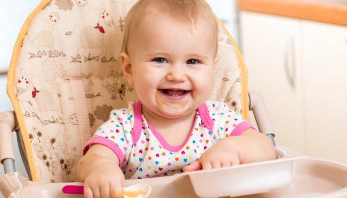 A baby girl sitting in a high chair and feeding herself with a baby spoon.