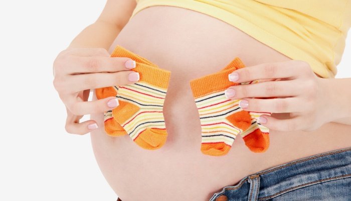 A woman holding two pairs of baby socks up to her pregnant belly.