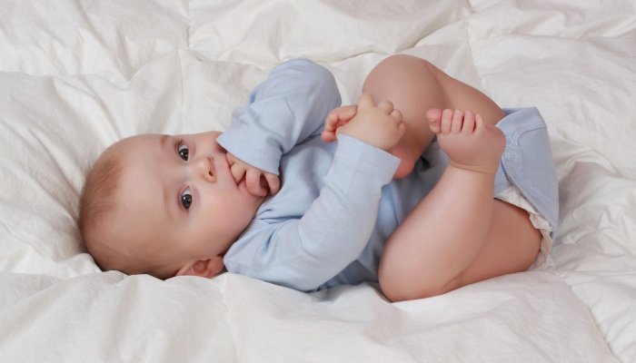 A cute baby boy on his back holding his foot and sucking on his fingers and bottom lip.