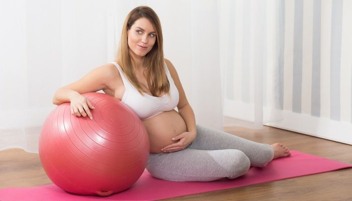 A pregnant lady sitting on a yoga mat with a pink exercise ball.