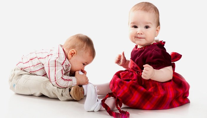 A baby boy biting his twin sister's shoe.