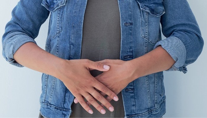 A woman in a jean jacket with her hands over her belly as if uncomfortable or in pain.
