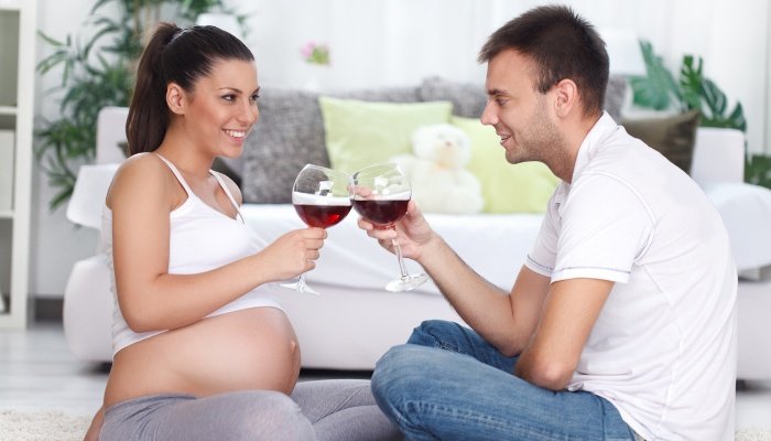A pregnant couple sitting on the floor toasting with glasses of wine.