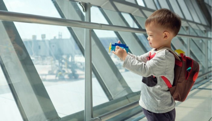 A cute toddler boy with a toy and a backpack standing at the windows of an airport.