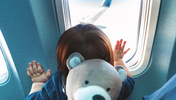 A toddler wearing a teddy bear backpack gazes out the window of an airplane.