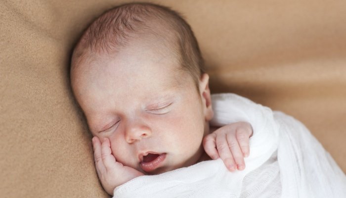 A swaddled newborn baby sleeping with his mouth open on a beige sheet.