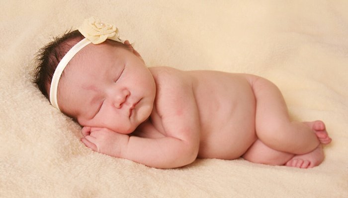 A newborn baby girl wearing nothing but a headband while lying on a peach blanket.