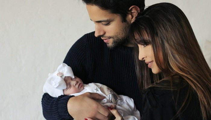 Parents dressed in black holding their newborn who is dressed in white with a white bow on her head.