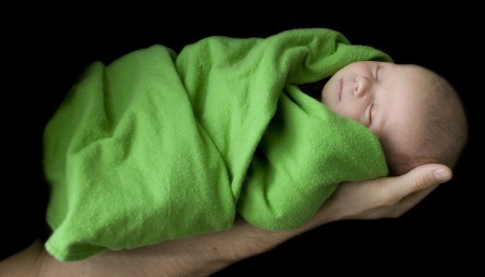 A newborn wrapped in a green blanket being held aloft on dad's arm against a black background.