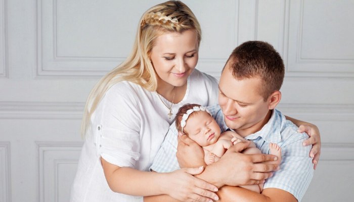 A new father sits in a rocking chair holding his newborn while the mother hugs them both.