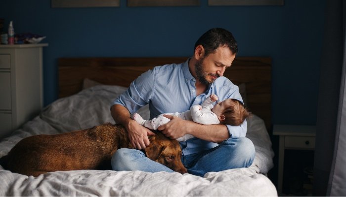 A dad sitting on the bed holding his newborn baby with his dog by his side.