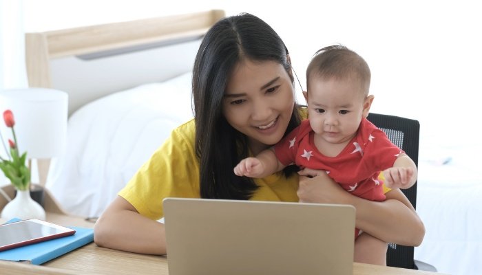 A young mom holding her baby while she smiles and works on her laptop.