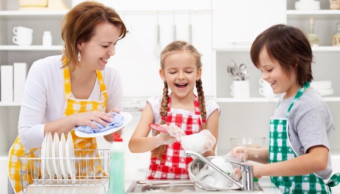 A mother and two children wear matching aprons while washing dishes together.