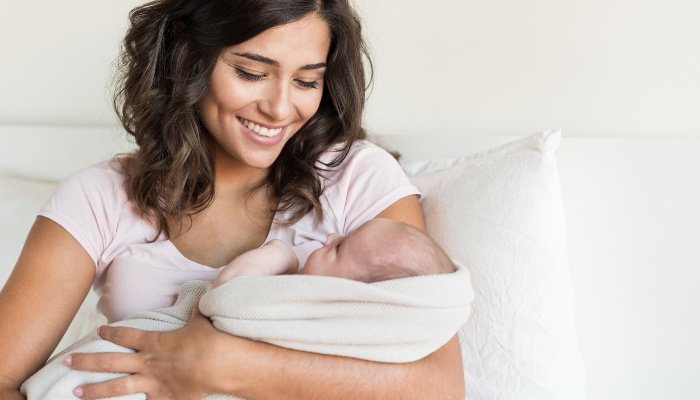 A young mother holding her newborn baby wrapped in a white blanket.