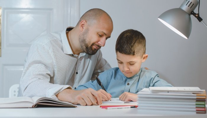 A father homeschooling his young son at a desk in a bedroom.